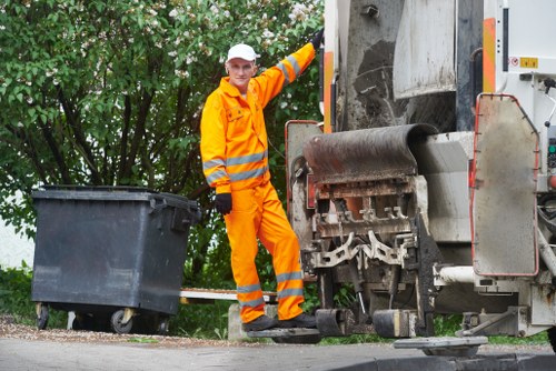 A male worker dressed in bright orange high-visibility safety clothing, including a jacket and trousers with reflective stripes, standing beside a large industrial rubbish collection vehicle. The worker is wearing a white hard hat and black gloves, and is positioned near an open waste hopper, which appears to be part of the refuse truck. In front of him, a large black plastic wheelie bin is placed on the pavement, ready for collection or emptying. The scene is set outdoors on a paved area, with a background of green leafy trees providing natural surroundings. The image captures the process of rubbish collection, emphasizing efficient waste management services that could be associated with companies like Garden Clearance Acton operating in the local area, including postcode W3 or W4 in west London. The lighting suggests daytime with natural sunlight illuminating the scene, highlighting the textures of the bin's smooth plastic surface, the metallic finish of the refuse vehicle, and the reflective strips on the worker's outfit.