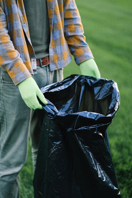 The image depicts two large, black plastic garbage bags filled with waste, placed on a paved sidewalk near a black metal fence with vertical bars. The bags appear to contain household rubbish, possibl