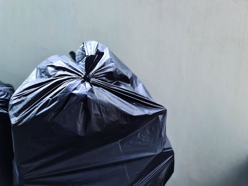 Close-up of a black plastic rubbish bag filled with waste, tightly knotted at the top, with a smooth, slightly shiny surface that has minor creases and folds. The bag is situated against a plain, ligh