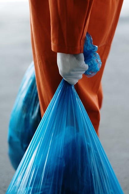 Close-up of a black plastic rubbish bag filled with waste, tightly knotted at the top, with a smooth, slightly shiny surface that has minor creases and folds. The bag is situated against a plain, ligh