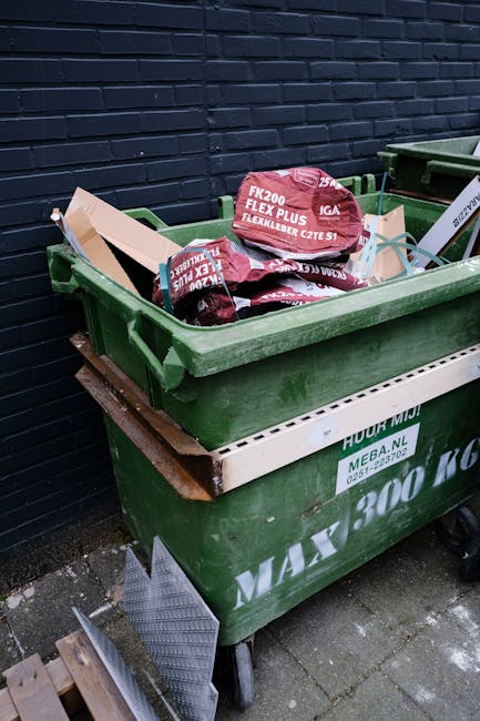 A large green wheeled refuse container positioned on a paved outdoor surface and situated next to a dark brick wall. Inside the container, there are multiple red bags of garden or landscaping material