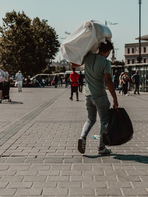 A young man is walking through an open urban square during daytime, carrying a large, white plastic bag filled with rubbish over his right shoulder and holding a black trash bag in his left hand. The 