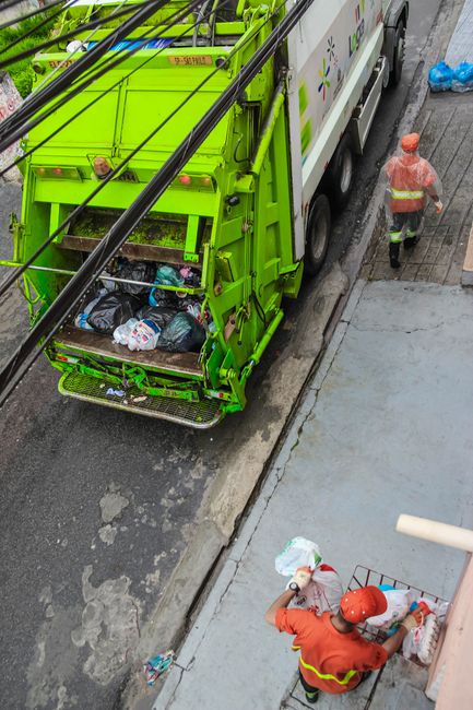 A roadside scene featuring a red garbage collection truck with its rear compactor open, positioned on the right side of the road. In front of the truck, a worker wearing a high-visibility orange vest 